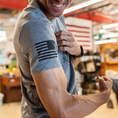 A man flexes his arm with a smile, wearing the Join or Die Tee - Tri-Blend, a gray shirt featuring a black American flag design on the sleeve inspired by Ben Franklin’s iconic “Join or Die” cartoon symbolizing colonial unity.