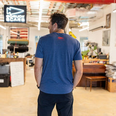 A man wearing the Graphic T-Shirt - 100% American stands indoors with his back to the camera in a cluttered, industrial-style room.