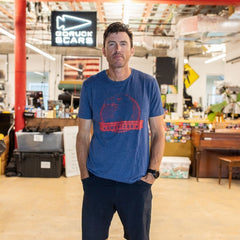 A man wearing the Graphic T-Shirt - 100% American stands in a casual, colorful office space with a “GORUCK SCARS” sign behind him.