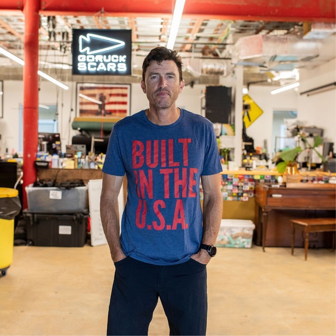 A man stands in a workshop wearing the Graphic T-Shirt - Built in the USA, featuring bold red text that reads “BUILT IN THE U.S.A.” on a blue background.