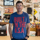 A man stands in a workshop wearing the Graphic T-Shirt - Built in the USA, featuring bold red text that reads “BUILT IN THE U.S.A.” on a blue background.