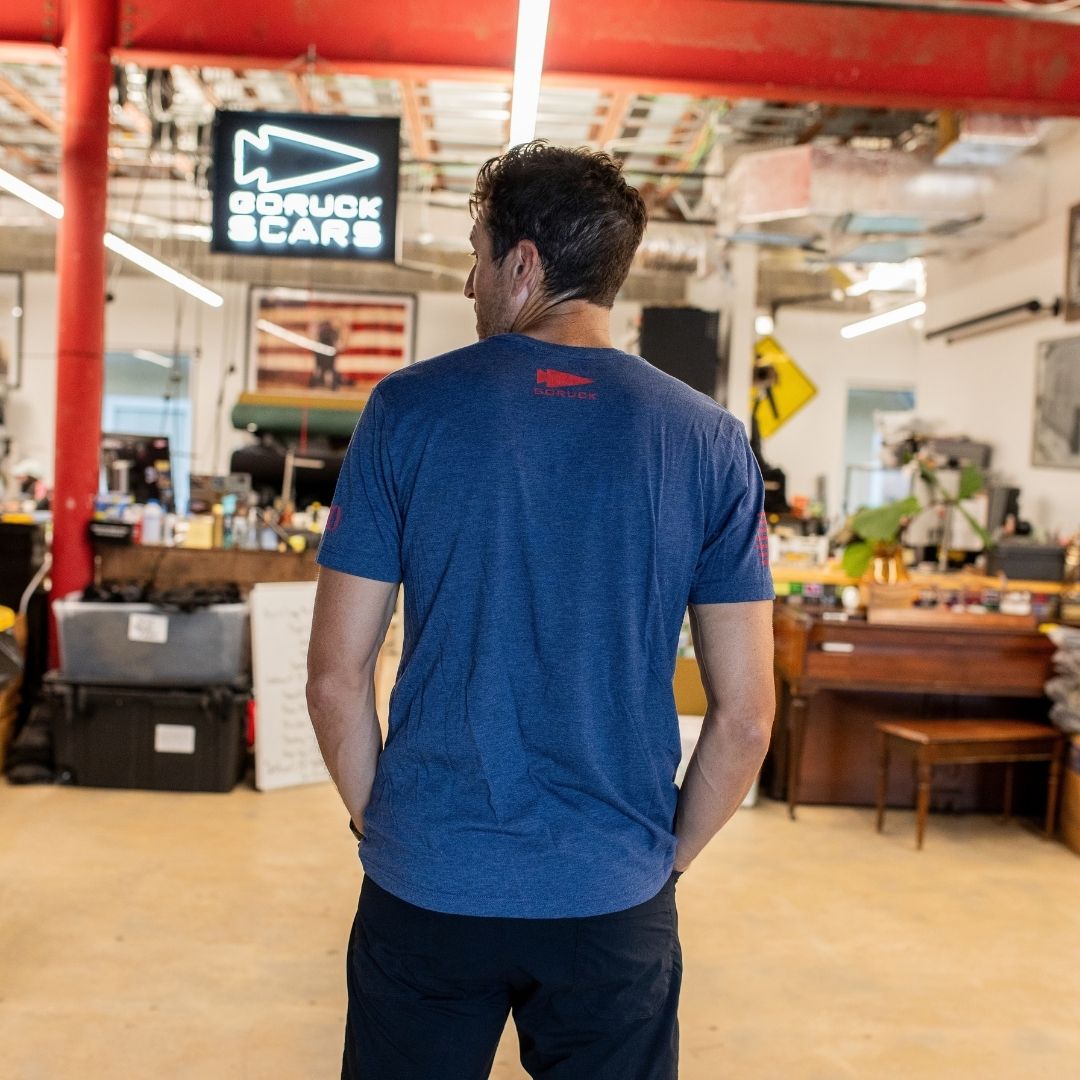 A man in the Join or Die Tee - Tri-Blend stands with his back to the camera in a cluttered, industrial-style workspace, evoking Ben Franklin’s inventive spirit.