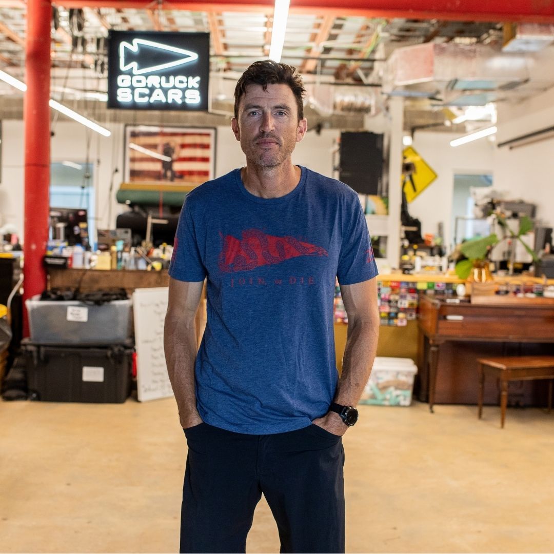 A man in a blue Join or Die Tee - Tri-Blend stands in an office with a “GORUCK SCARS” sign, desks, and an American flag behind him, evoking the spirit of colonial unity seen in Ben Franklin’s famous “Join or Die” cartoon.