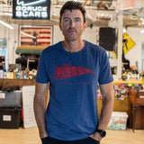 A man in a blue Join or Die Tee - Tri-Blend stands in an office with a “GORUCK SCARS” sign, desks, and an American flag behind him, evoking the spirit of colonial unity seen in Ben Franklin’s famous “Join or Die” cartoon.