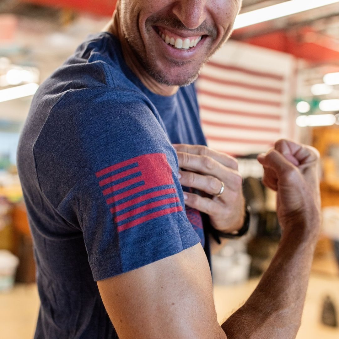 A smiling person flexes their arm, revealing a red American flag motif on the sleeve of the Join or Die Tee - Tri-Blend in blue, echoing the colonial unity symbolized in Ben Franklin’s iconic “Join or Die” cartoon.