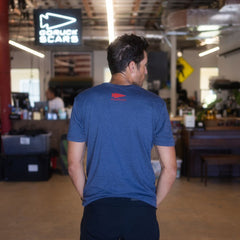 A man stands indoors with his back to the camera in a casual, cluttered workspace, wearing the DFQ TEE - TRI-BLENDX in blue. The motivational “Don’t F*ing Quit” message is displayed on the shirt, highlighting both its comfort and inspiring design.