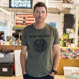 A man stands in an office wearing the “In GOD We Trust” t-shirt and shorts, looking at the camera.