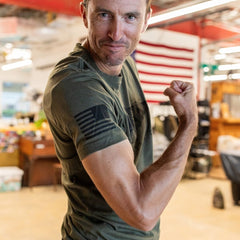 Wearing the Join or Die Tee - Tri-Blend, a man in a green shirt flexes his arm and smiles against the backdrop of an American flag, capturing the spirit of colonial unity inspired by Ben Franklin’s iconic “Join or Die” cartoon.