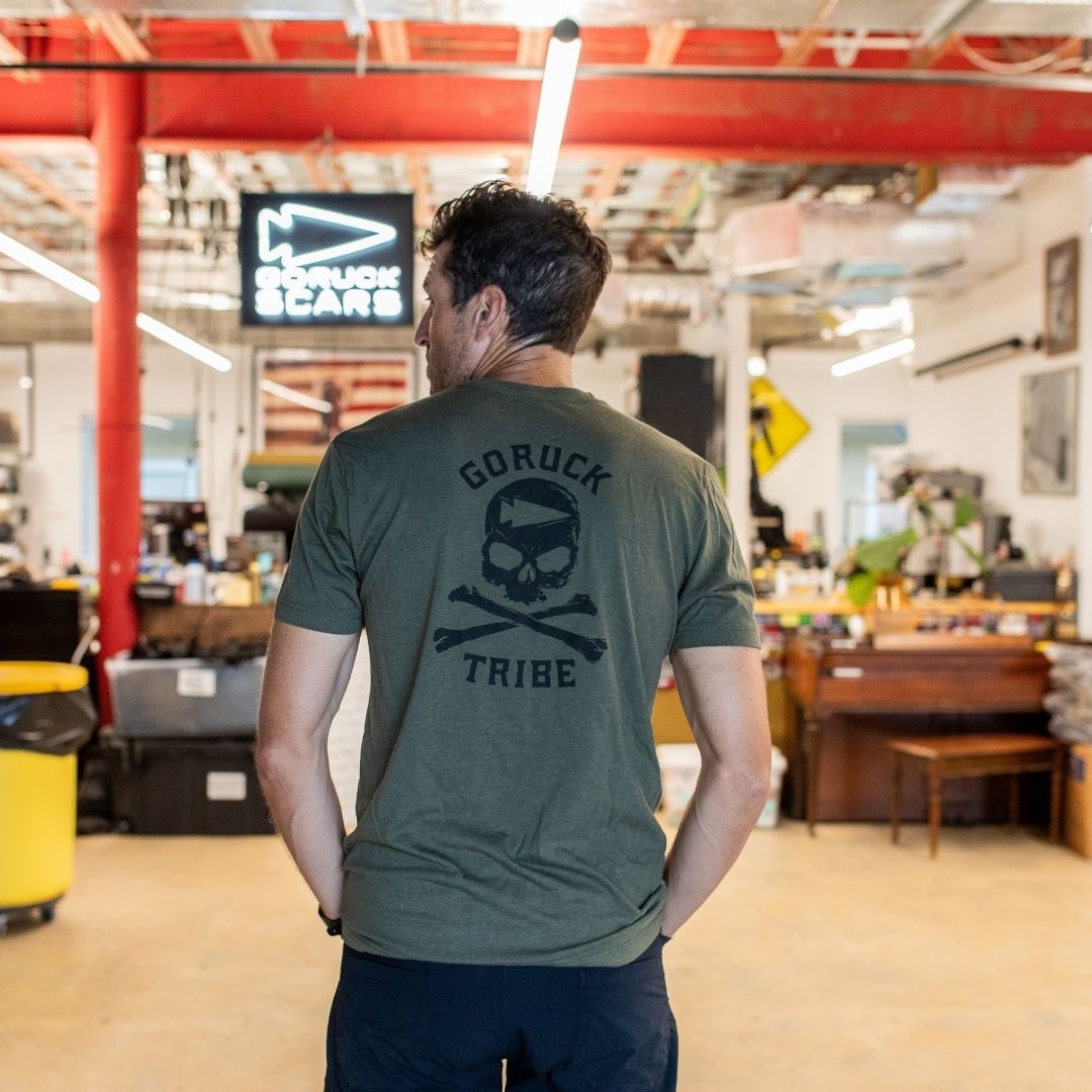 A man stands with his back turned in an office space, wearing the olive “Join or Die Tee - Tri-Blend” featuring a skull and crossbones—a design inspired by colonial unity and Ben Franklin’s legendary Join or Die cartoon.