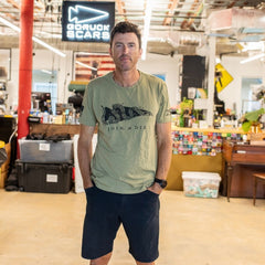 A man wearing the Join or Die Tee - Tri-Blend and navy shorts stands in an office with a “GORUCK SCARS” sign in the background, evoking the spirit of Ben Franklin’s iconic “Join or Die” cartoon and its call for colonial unity.