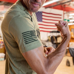 A man flexes his arm, wearing the Join or Die Tee – Tri-Blend in green, featuring a US flag design. The American flag is blurred in the background, capturing strength and patriotism inspired by Ben Franklin’s iconic message of colonial unity from the “Join or Die” cartoon.