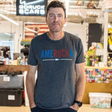 Man standing in warehouse, wearing “AmeRUCKa” t-shirt, with hands in pockets, smiling at camera.