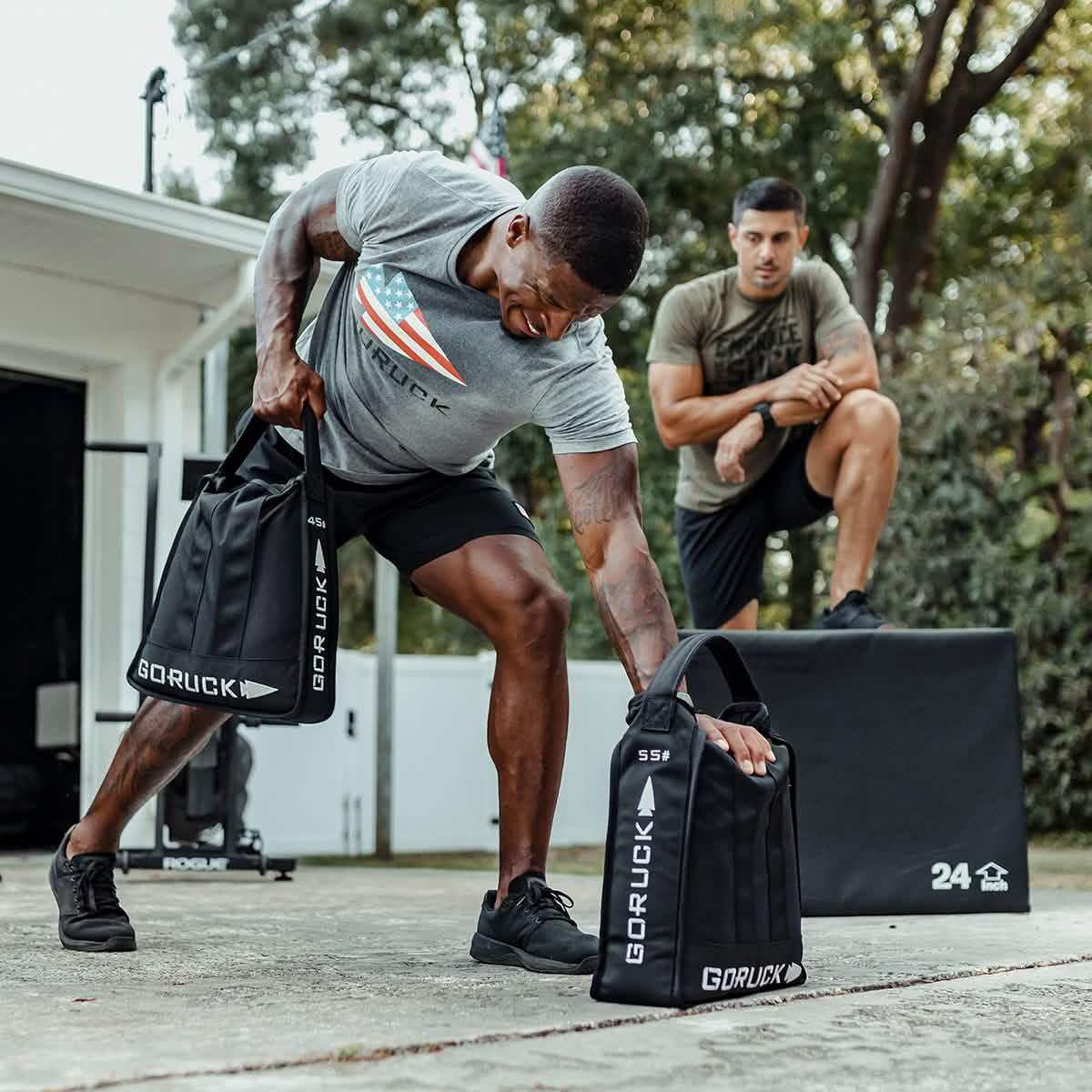 Man lifting GORUCK sandbag outdoors, another man watches, rucking gear in fitness training
