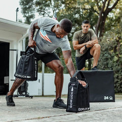 Man lifting GORUCK sandbag outdoors, another man watches, rucking gear in fitness training