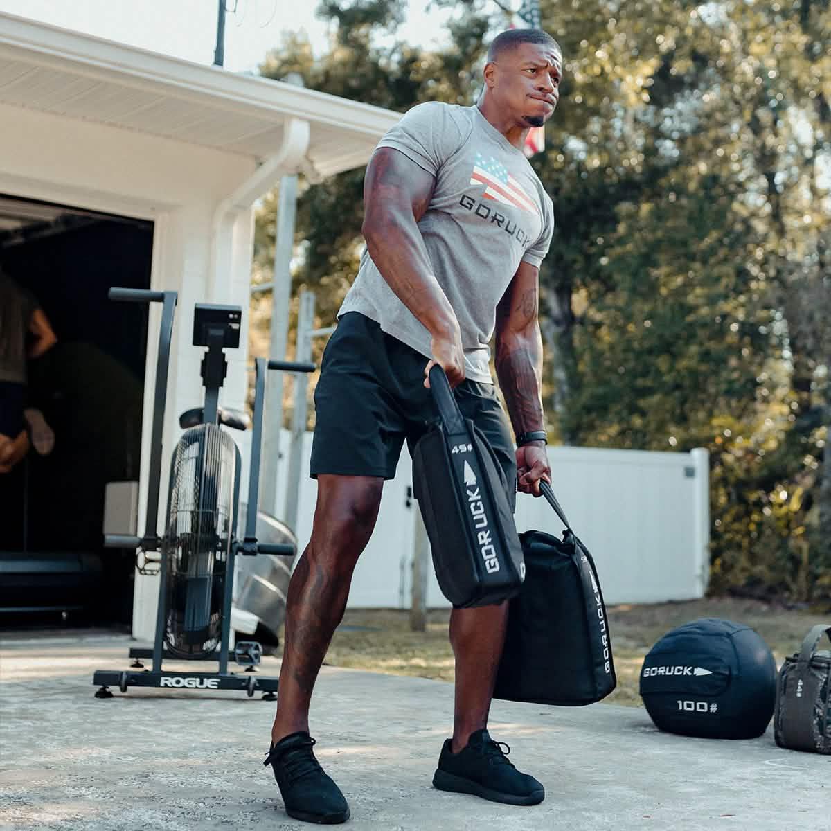 Man lifting GORUCK training sandbags outdoors, wearing GORUCK shirt and athletic gear