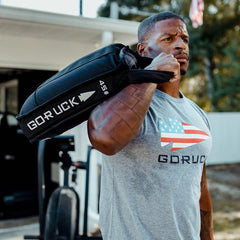 Man training outdoors with GORUCK 45 lb sandbag, wearing GORUCK logo shirt