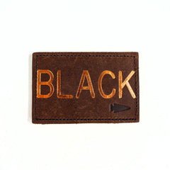 The Patch - Custom Leather Monogram is a brown leather patch, personalized with "BLACK" embossed in gold and accented with a small arrow symbol in the corner.