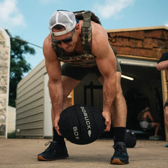Man lifting GORUCK 60LB medicine ball outdoors, wearing tactical gear and cap
