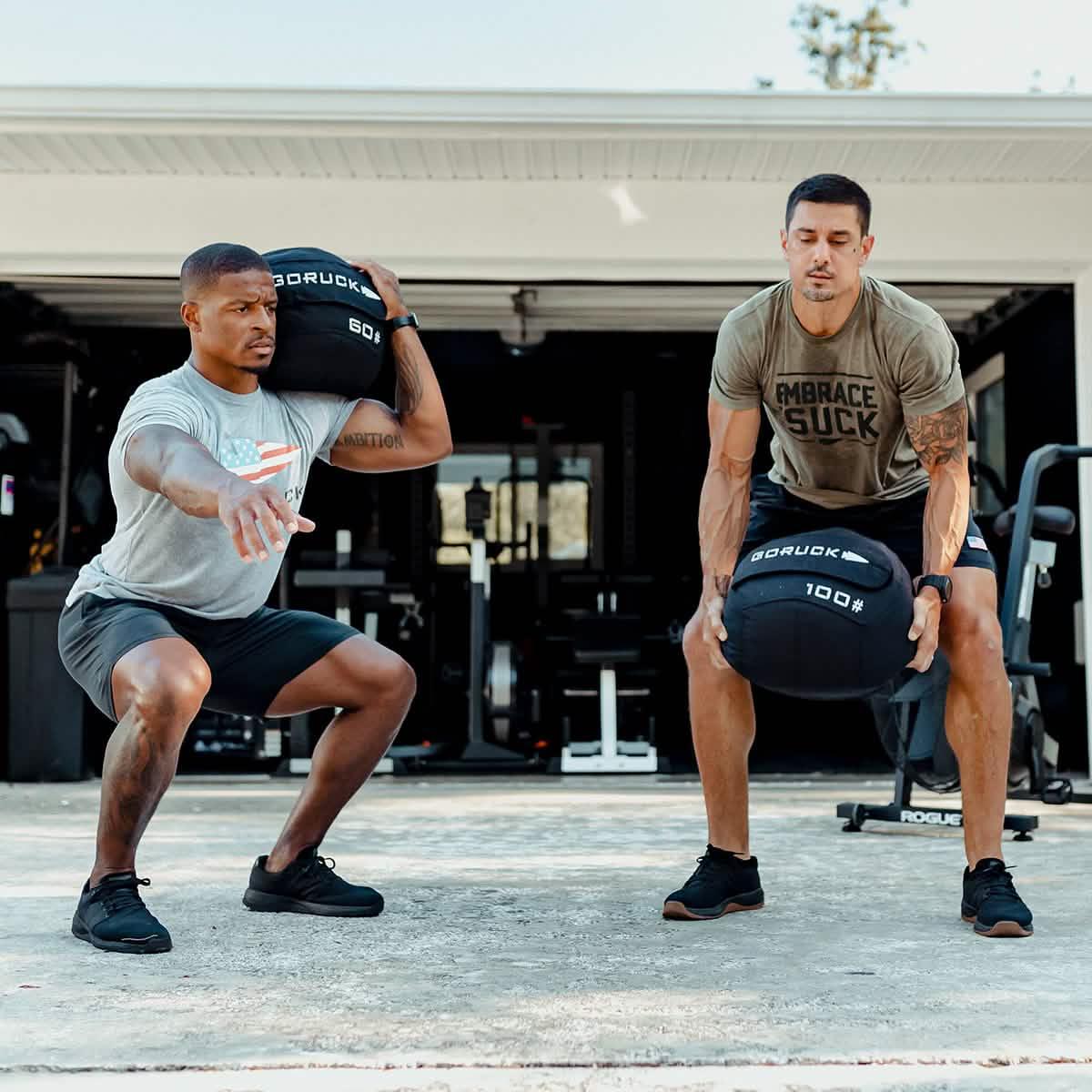 Two men rucking outside a garage gym using GORUCK weighted sandbags for strength training.