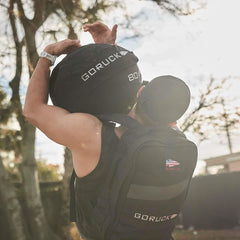 Man outdoors lifting GORUCK 80lb sandbag, wearing GORUCK backpack, fitness training gear