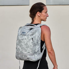 A woman with sunburned shoulders stands against a light wall, wearing the GR0 - Ripstop ROBIC® gray water-resistant backpack.