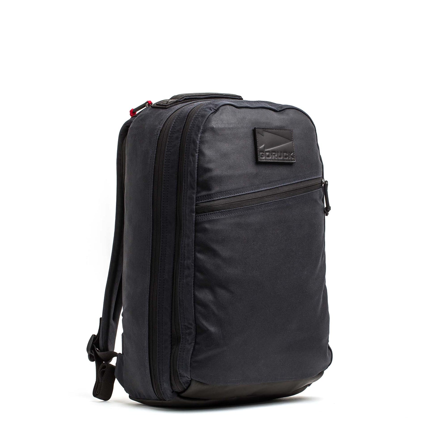 The Double Compartment Bullet Ruck - Heritage Waxed Canvas - 18L is a black backpack featuring a front zipper pocket and side buckle, displayed upright against a white background.