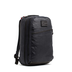 The Double Compartment Bullet Ruck - Heritage Waxed Canvas - 18L is a black backpack featuring a front zipper pocket and side buckle, displayed upright against a white background.
