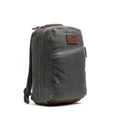 The Double Compartment Bullet Ruck - Heritage Waxed Canvas - 18L features dark gray waxed canvas, leather trim, dual zippers, and a brown front logo patch—making it a stylish and functional travel daypack.
