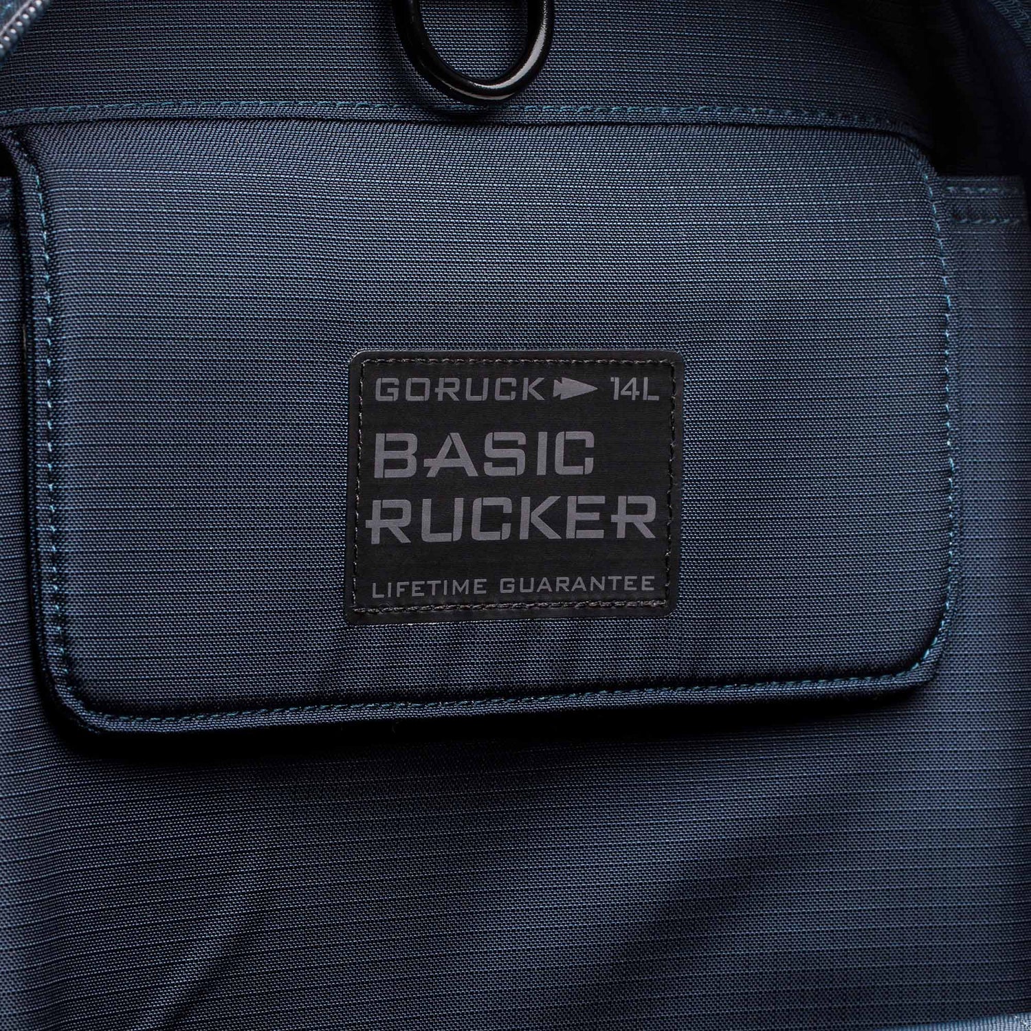 Close-up of the front pocket on a blue Basic Rucker backpack, featuring a stitched label and designed for comfortable rucking with secure storage for your ruck plate.