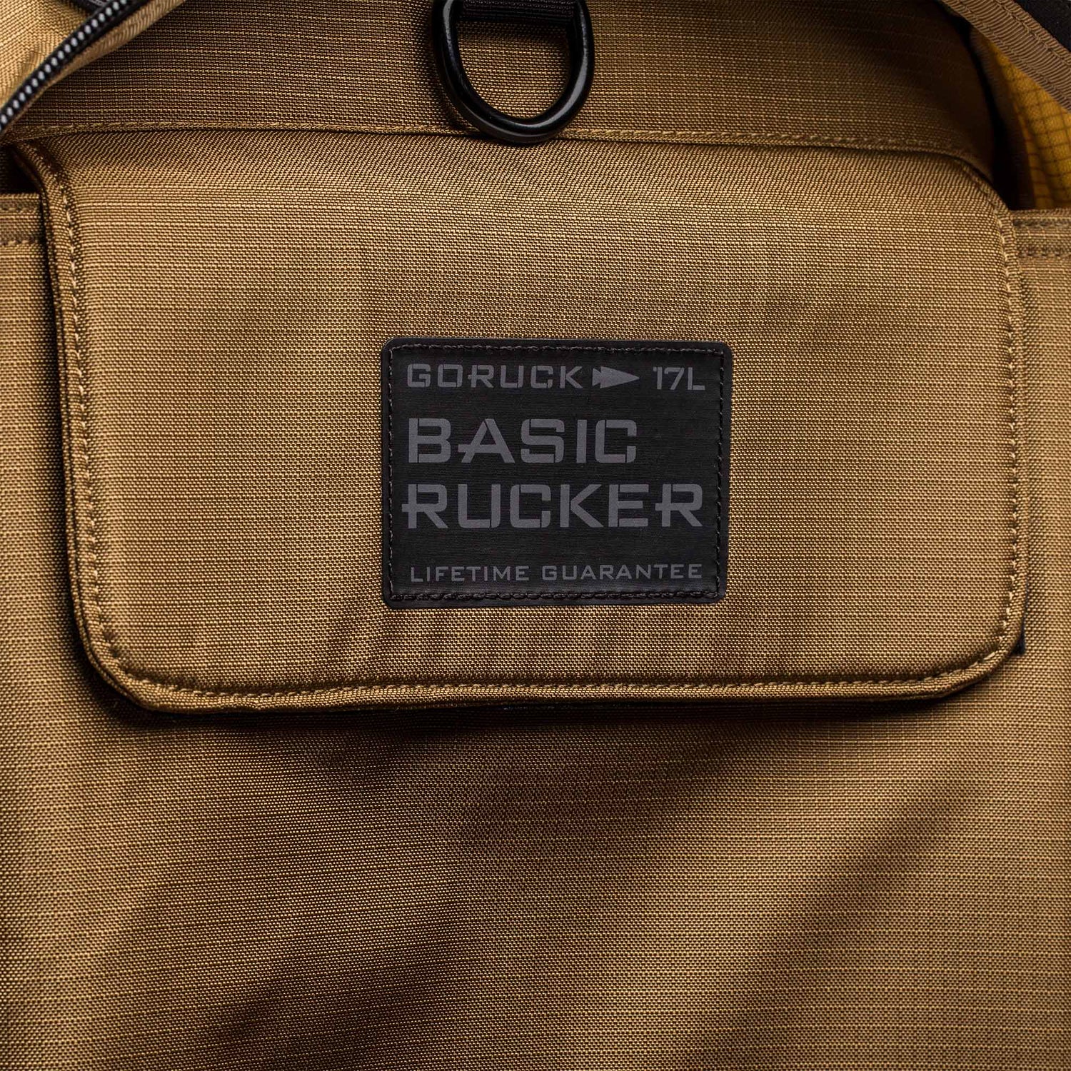 Close-up of a tan Basic Rucker® backpack pocket, featuring a label that reads 