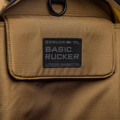 Close-up of a tan Basic Rucker® backpack pocket, featuring a label that reads "17L BASIC RUCKER LIFETIME GUARANTEE.