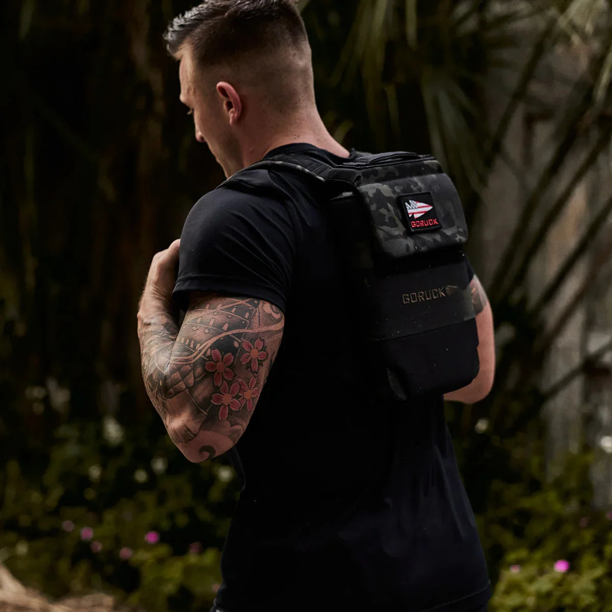 Man outdoors wearing black GORUCK rucksack with USA flag patch, showing tattooed arm
