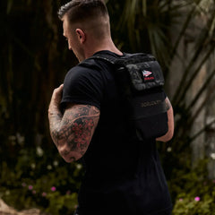 Man outdoors wearing black GORUCK rucksack with USA flag patch, showing tattooed arm