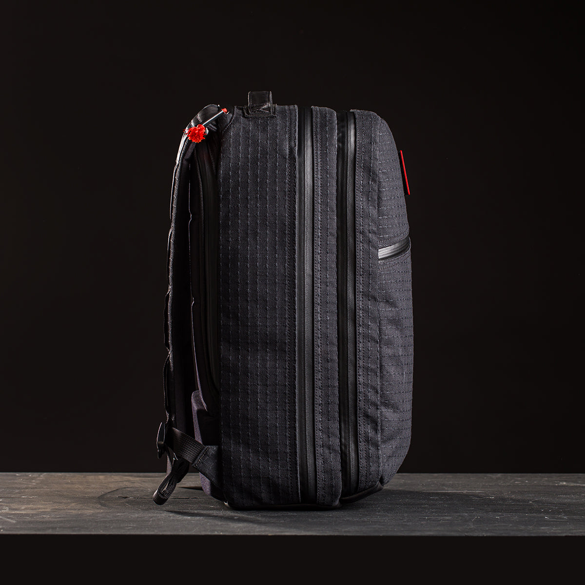 A limited-edition Bullet Ruck x Carryology - GRXC2 Samurai 2.0 backpack in black with vertical stitching, side zippers, and a red zipper pull, inspired by the JPXDP 17oz Black Samurai Sashiko, shown against a dark background.
