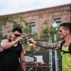 Two men in GORUCK gear cheers with camo can covers outdoors, urban backdrop