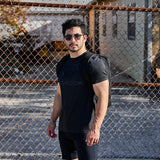 Man wearing sunglasses and a backpack with the GORUCK Spearhead Tee - Tri-BlendX stands in front of a chain-link fence on a sunny day.