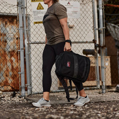 Woman in GORUCK black leggings and tee carrying black rucksack outdoors near chain-link fence