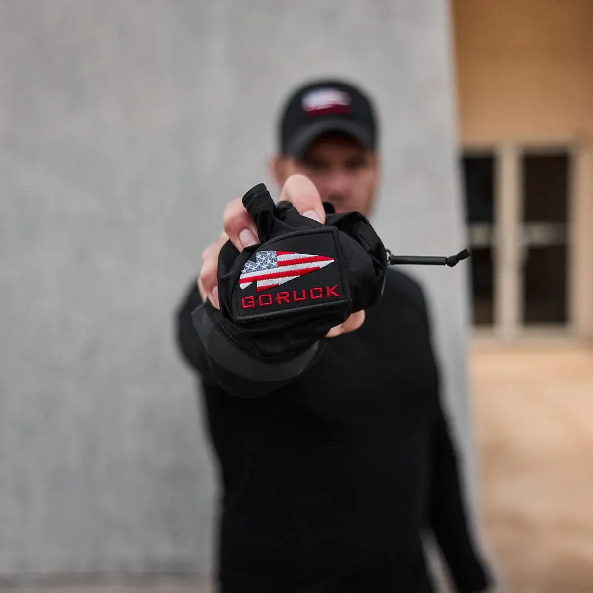 Man in black holds out a small black Packable Bullet Ruck - Ripstop ROBIC® with an American flag patch, ideal as a compact day bag; background is blurred.