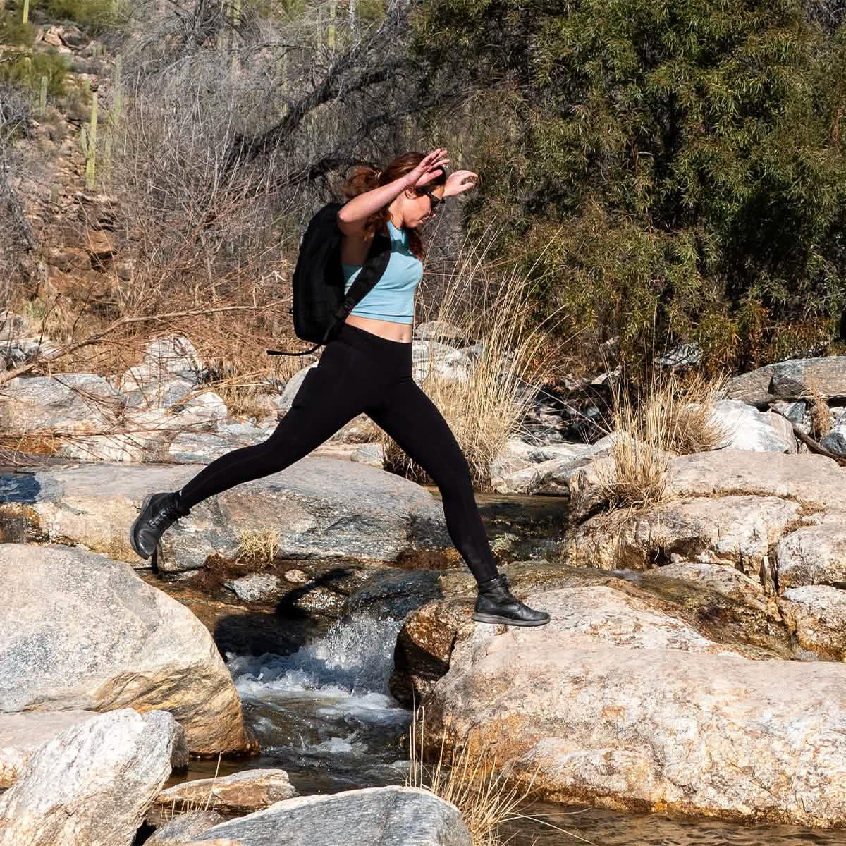 Woman rucking outdoors with GORUCK backpack, leaping over rocky creek in activewear