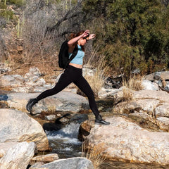Woman rucking outdoors with GORUCK backpack, leaping over rocky creek in activewear