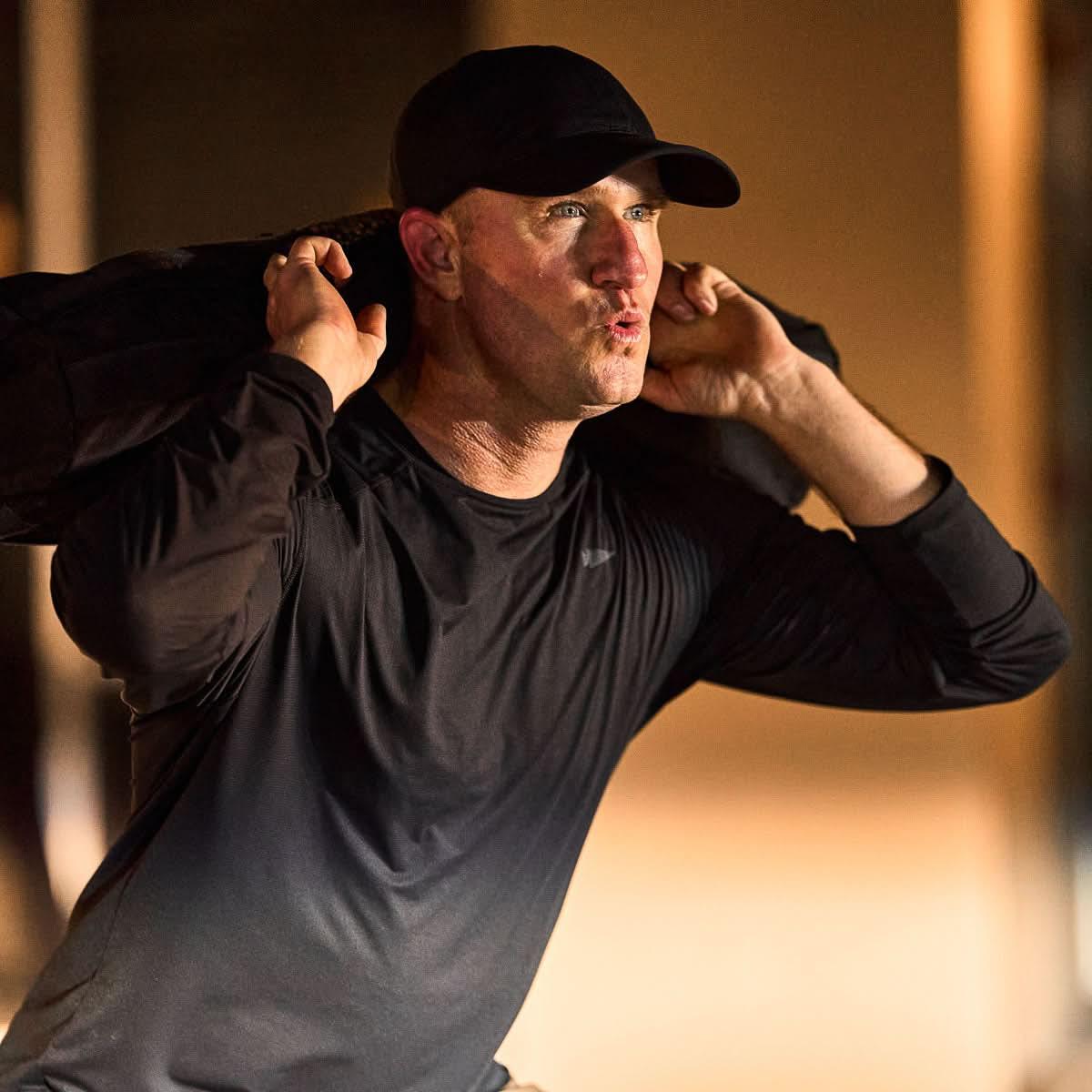 Man wearing GORUCK black running hat and shirt, lifting a weighted ruck indoors