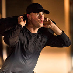 Man wearing GORUCK black running hat and shirt, lifting a weighted ruck indoors