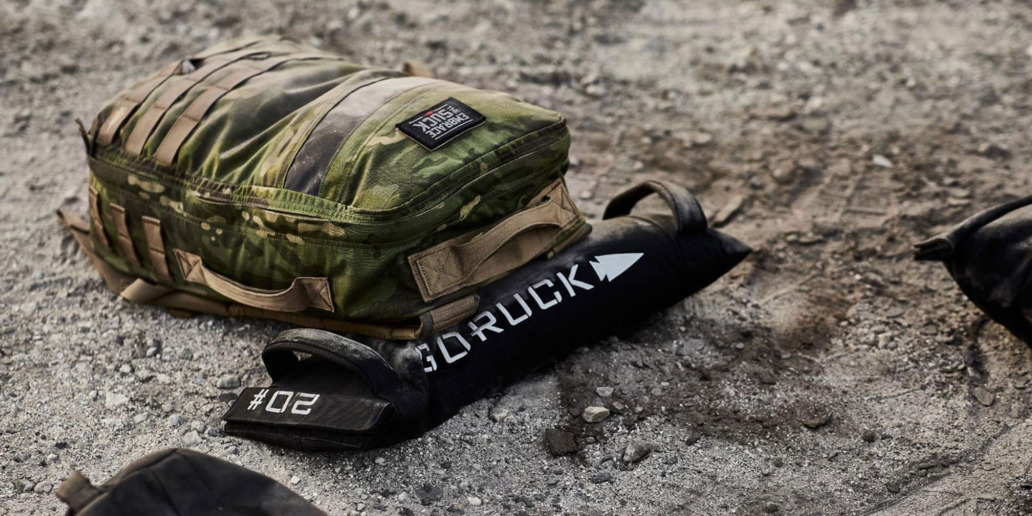 A camo backpack and a black GORUCK sandbag rest on gravel.