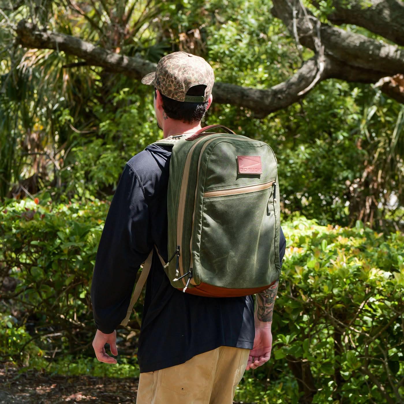 Man wearing a GORUCK olive green rucksack outdoors in a leafy, sunlit setting.