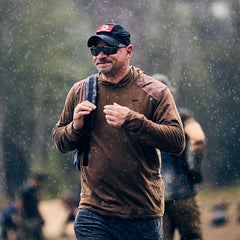Man wearing a cap and sunglasses walks in the rain with the Double Compartment Bullet Ruck - Ballistic Nylon Cordura - 18L, featuring a bombproof laptop compartment; blurred people appear in the background.