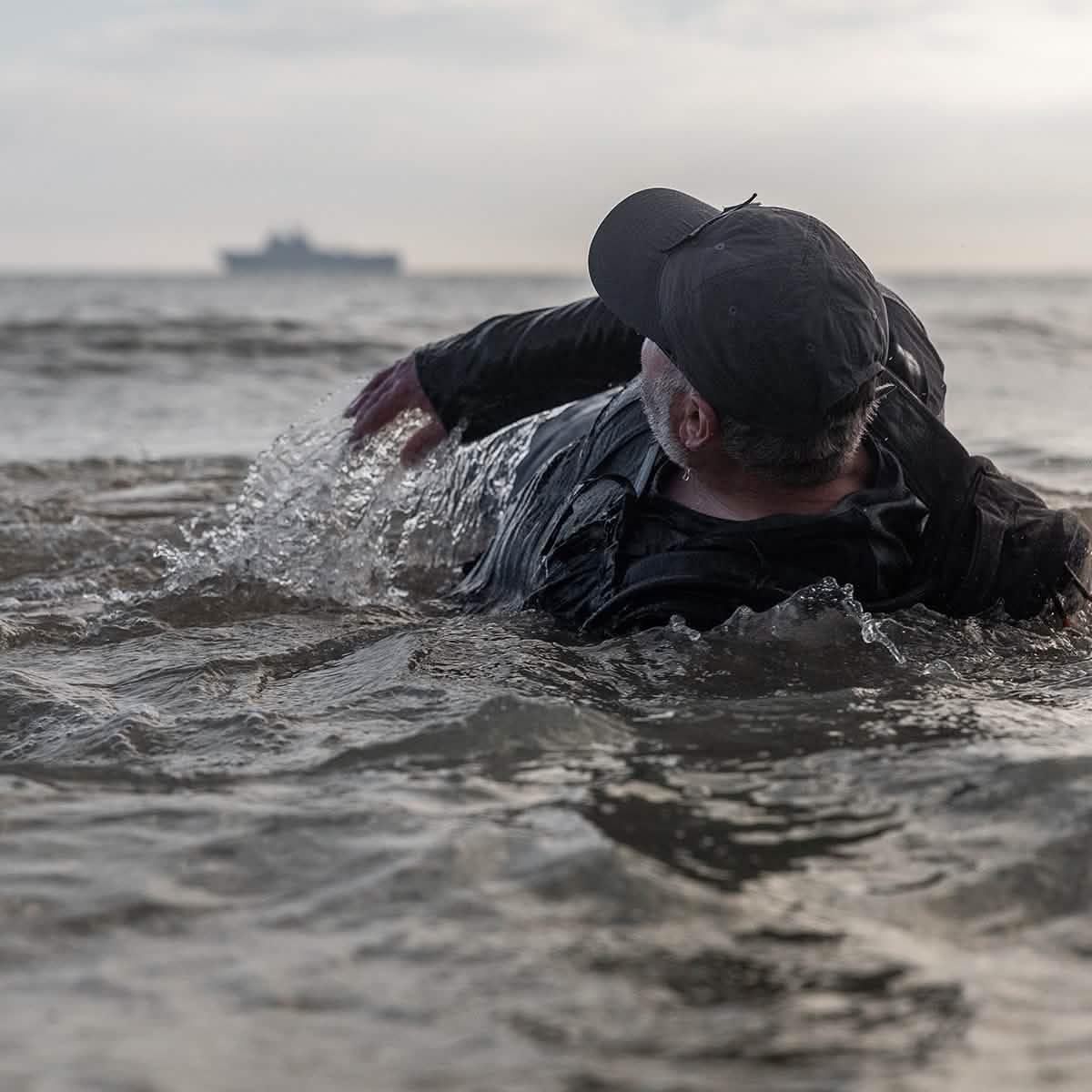 Man in GORUCK gear rucking through ocean surf, ship in background, rugged training scene