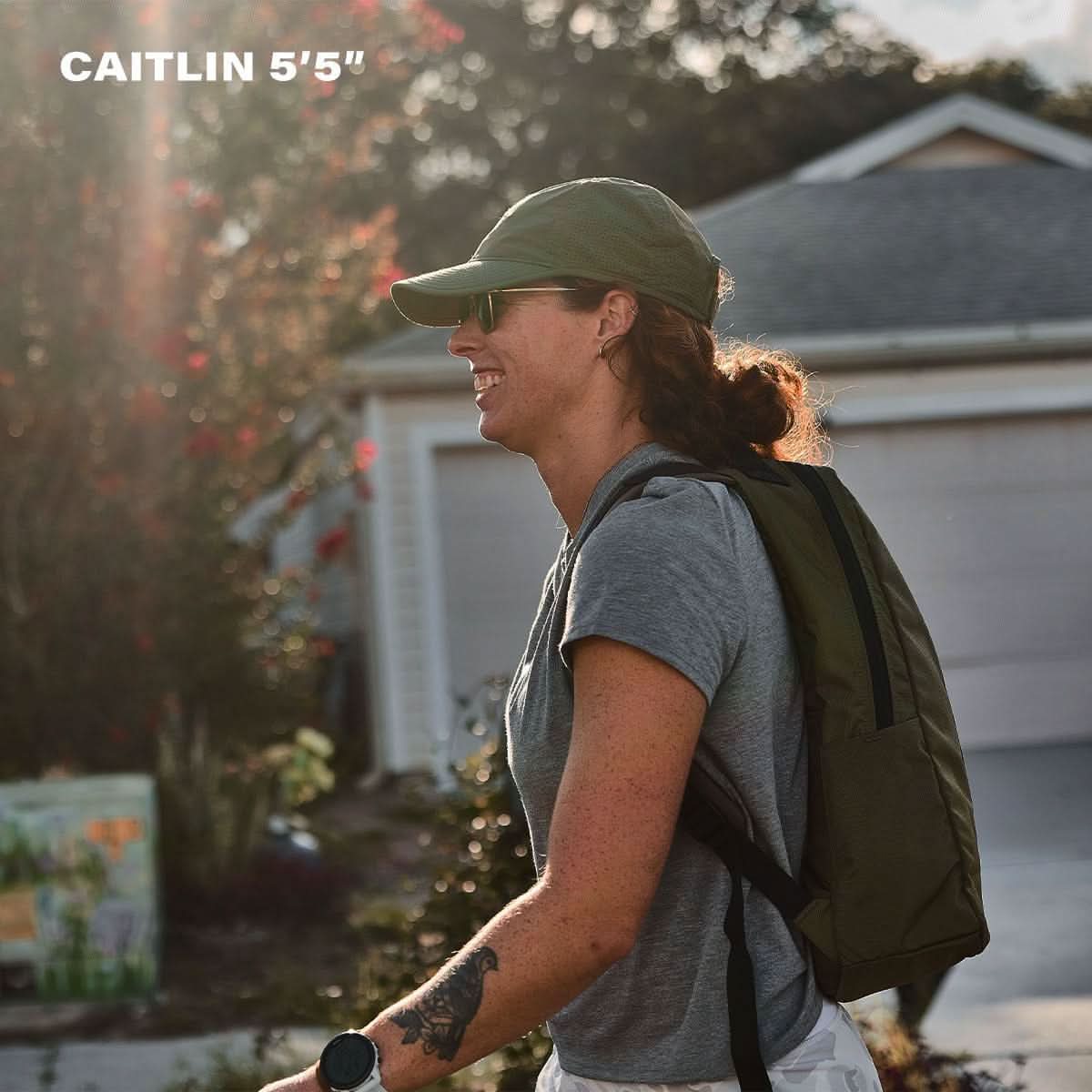 Woman rucking with GORUCK backpack, wearing cap, sunglasses, and gray shirt outdoors