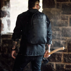 Man wearing black GORUCK backpack in rustic stone setting, holding an axe behind his back