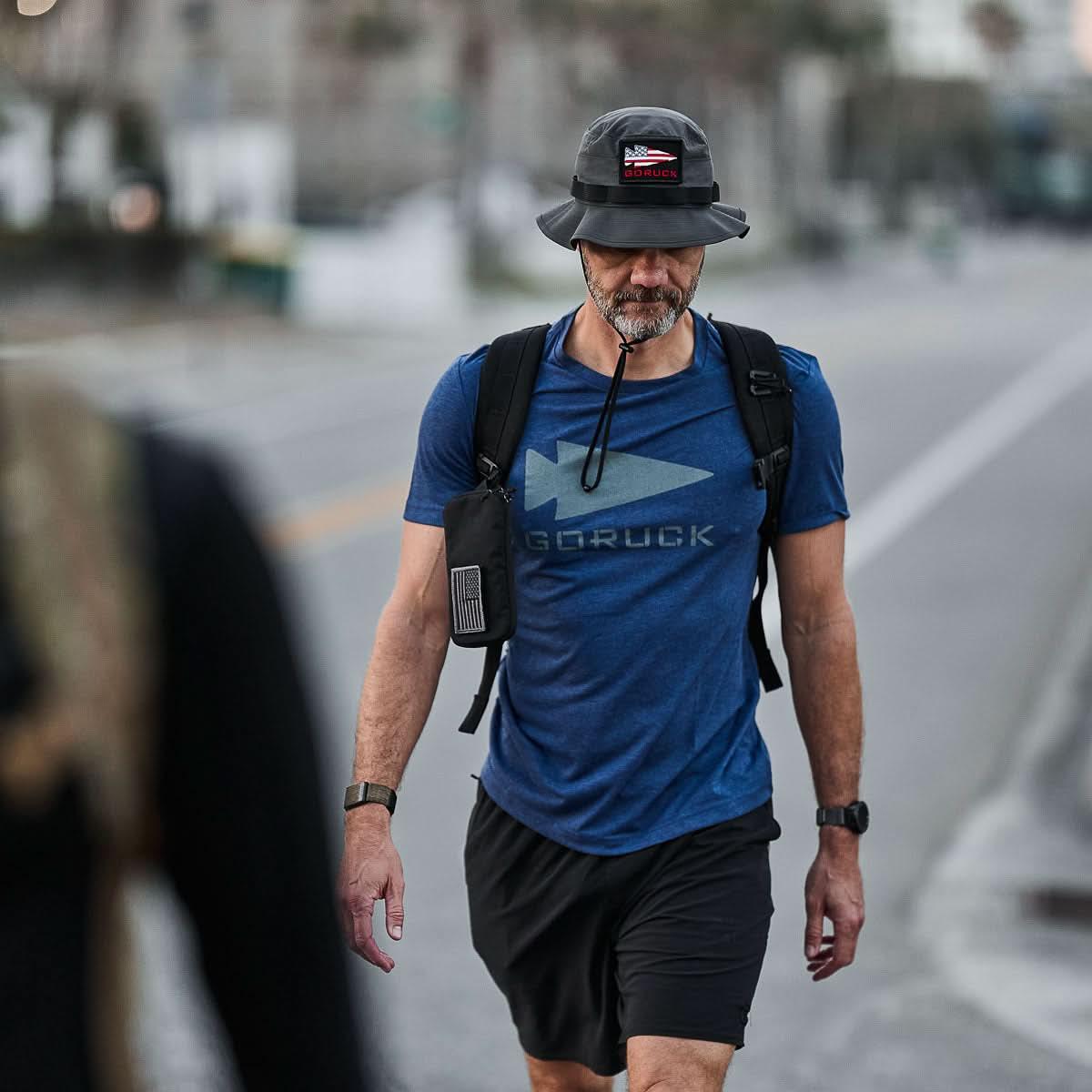 Man wearing GORUCK charcoal boonie hat, blue GORUCK shirt, backpack, rucking outdoors