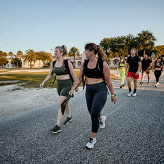 A group of people strolls and chats on a sunny road, framed by palm trees and buildings. The women wear Women's Cropped Training Leggings - ToughFlex, offering comfort and flexibility under the warm sun.